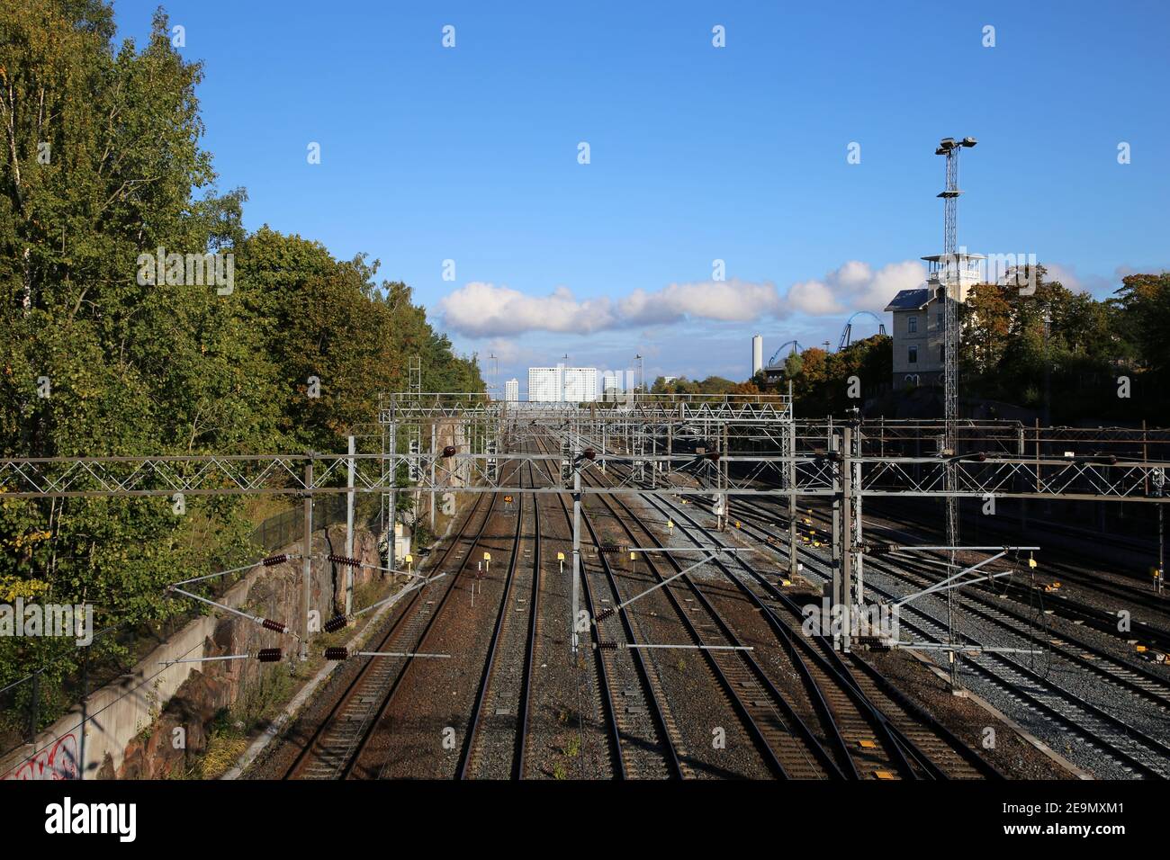 Railroad in Helsinki, Finland. Photographed Sep, 2019. Multiple train tracks with power supply lines, some trees, blue sky and the Mall of Tripla. Stock Photo