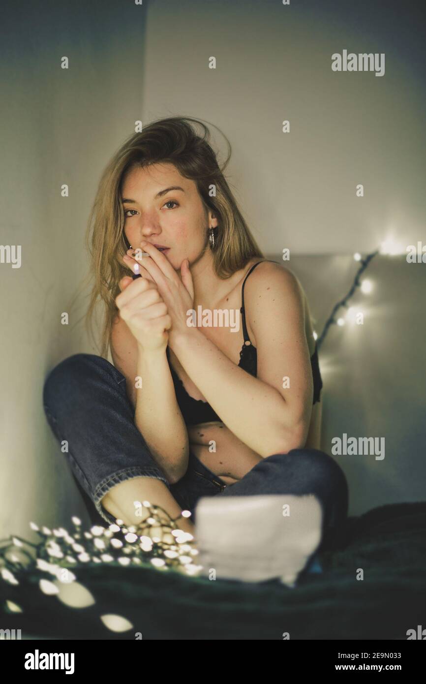 Vertical shot of a Caucasian female lighting a cigarette while sitting on her bed Stock Photo