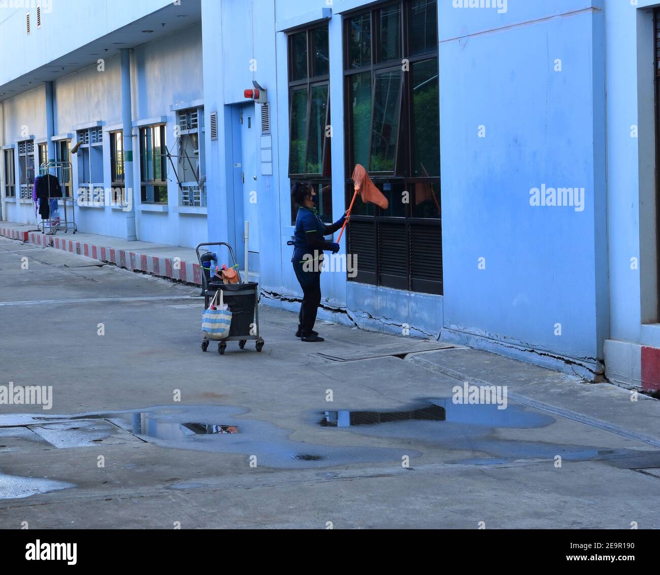 Bangkok, Thailand - February 2, 2021 : Blurred motion of female worker wearing face mask using mop with cloth to wipe glass window outside a building Stock Photo