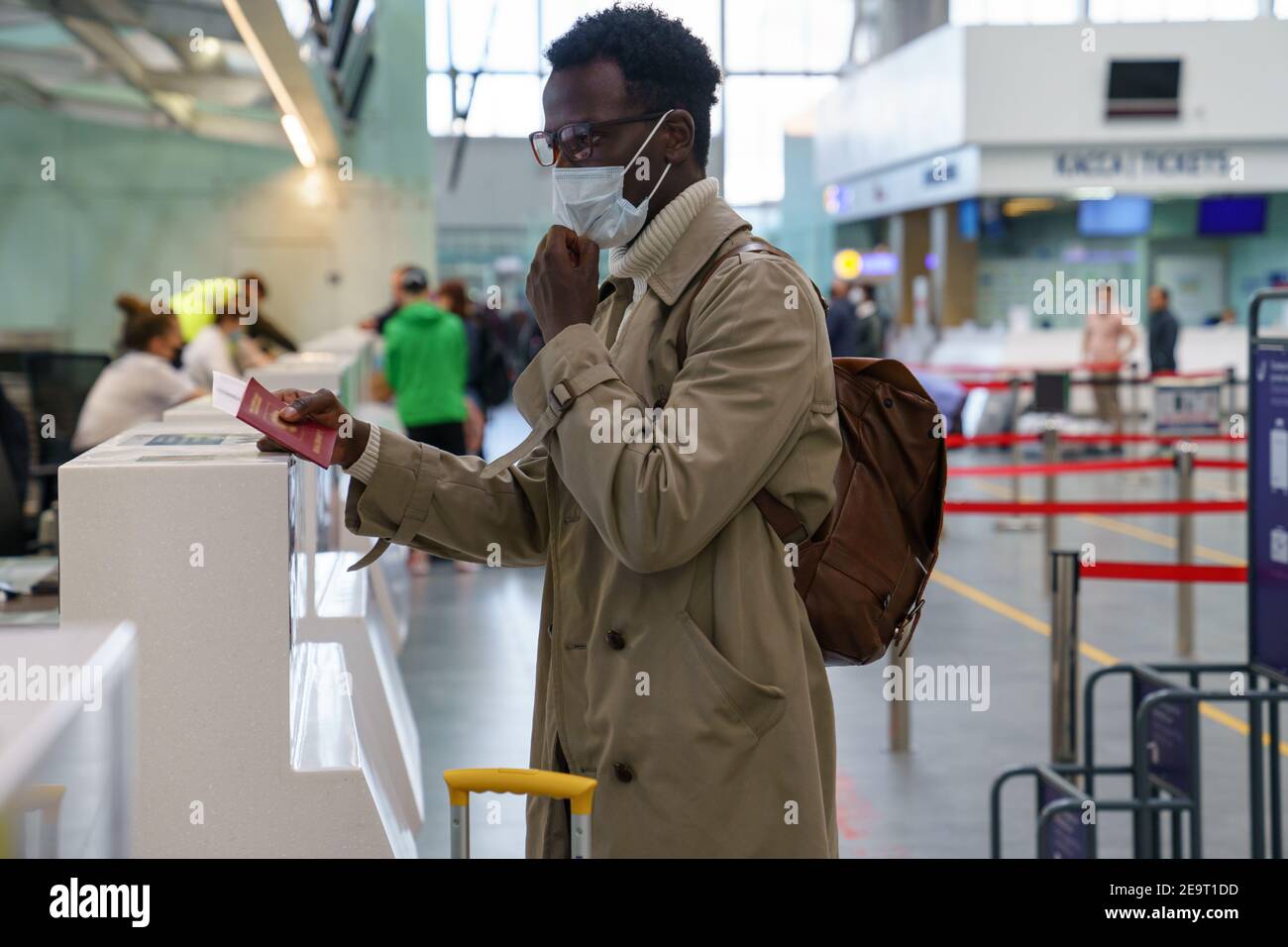Black man stands at check-in counters at the airport, giving passport to an officer, wear face mask. Stock Photo