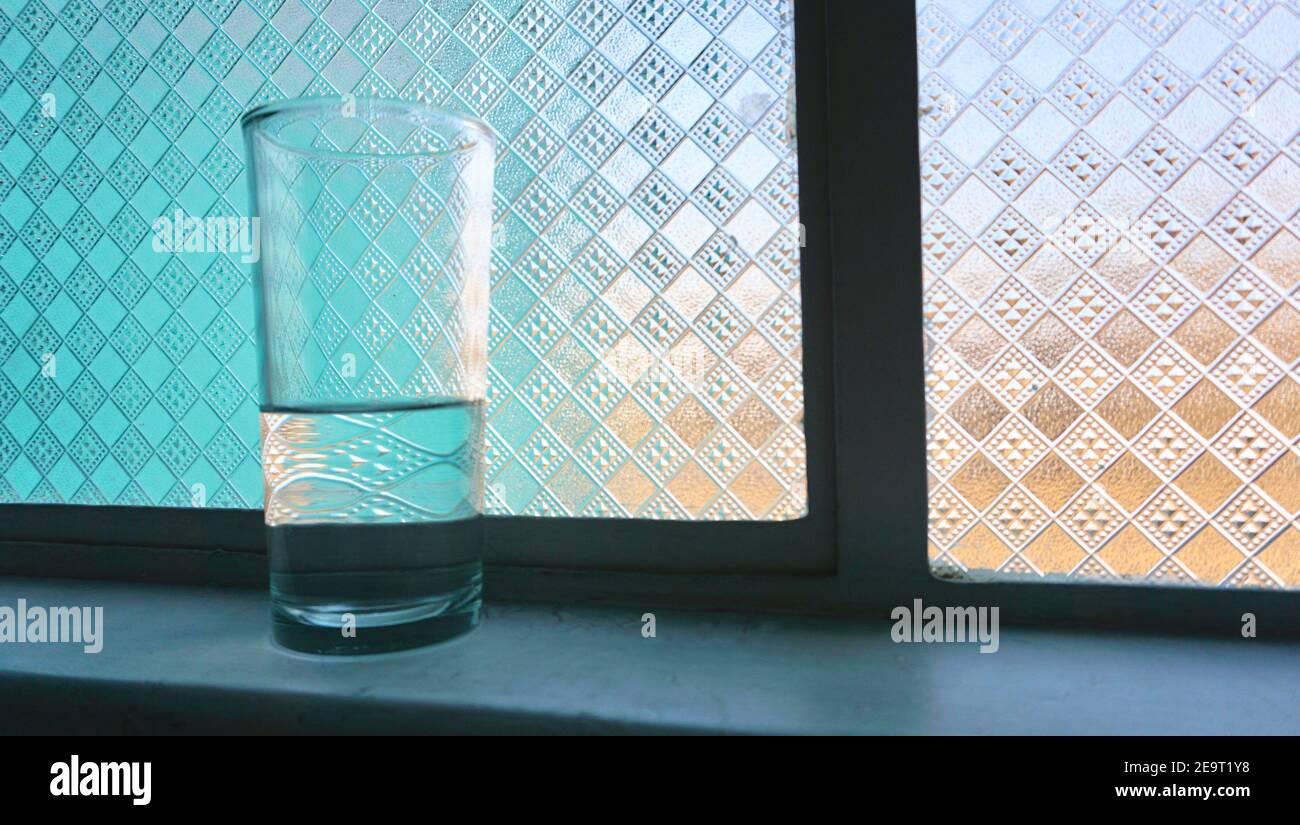 Glass of water less than half full on a window ledge with textured glass behind it. Stock Photo