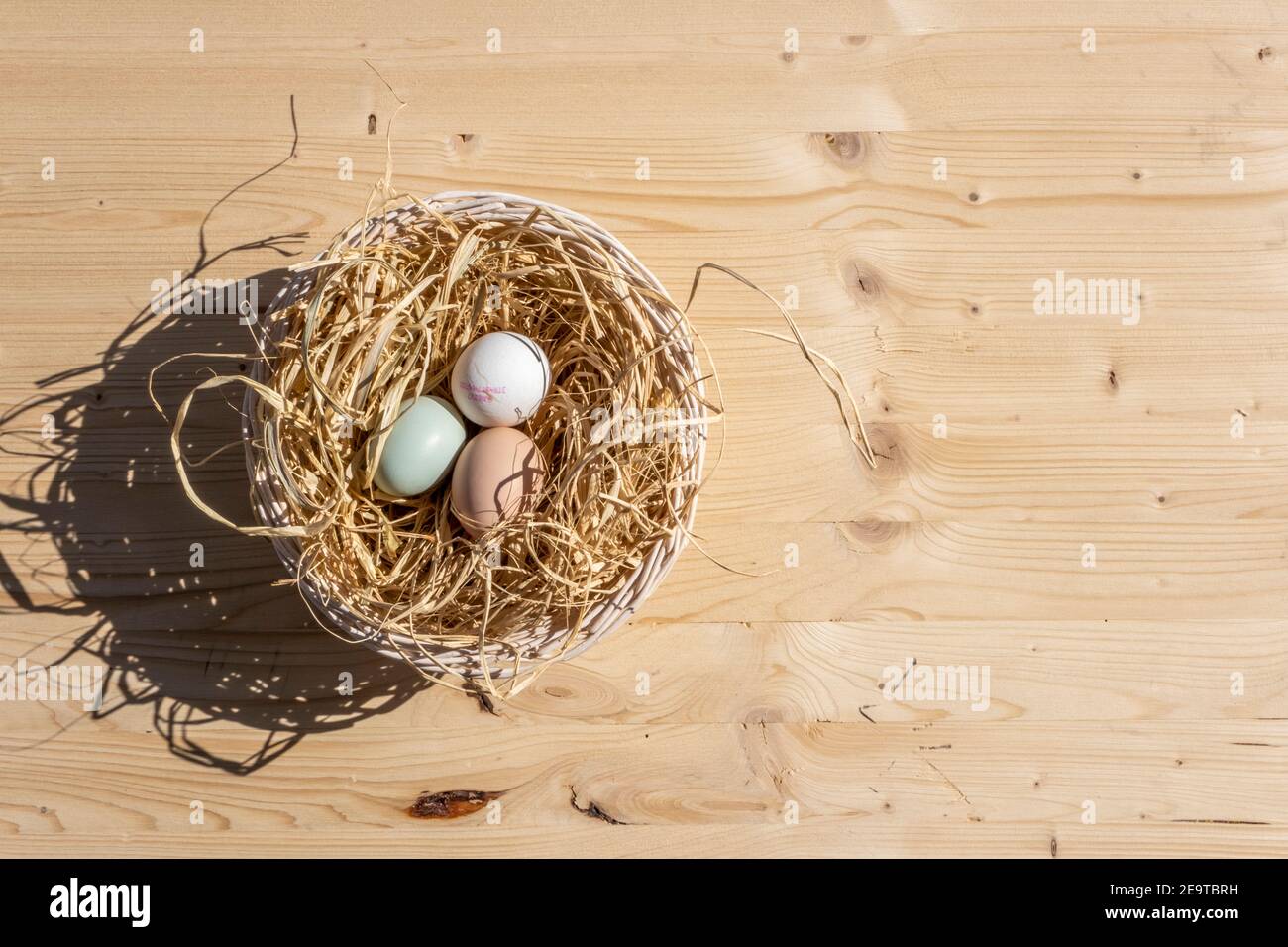 Organic Easter concept: Top view on a basket filled with straw and three natural colored eggs. Sun light creates shadow on wooden table surface. Stock Photo