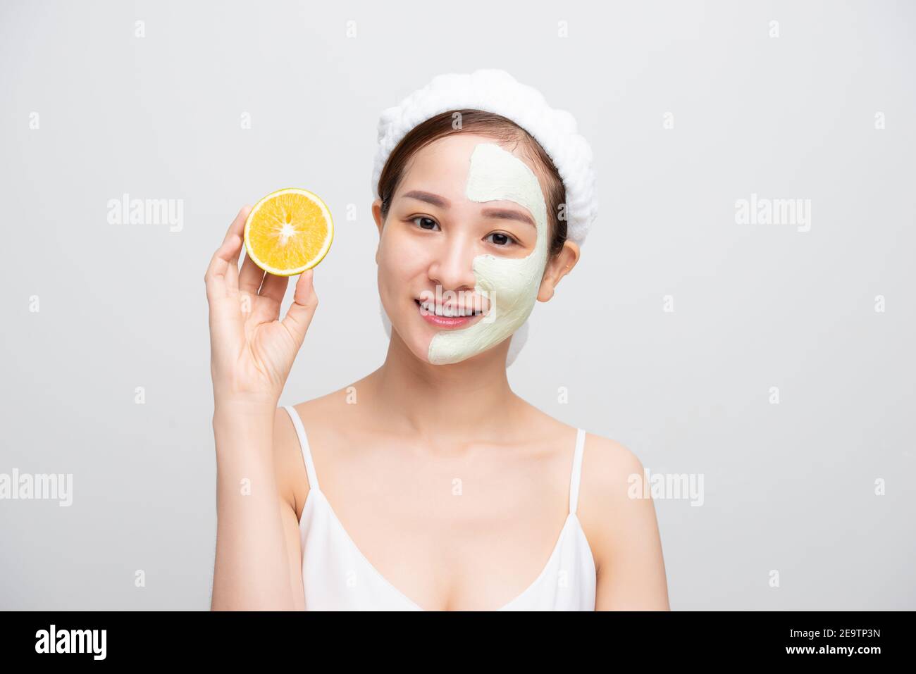 Beautiful woman is getting facial clay mask and holding orange pieces on white background. Beauty, body care and spa concept Stock Photo
