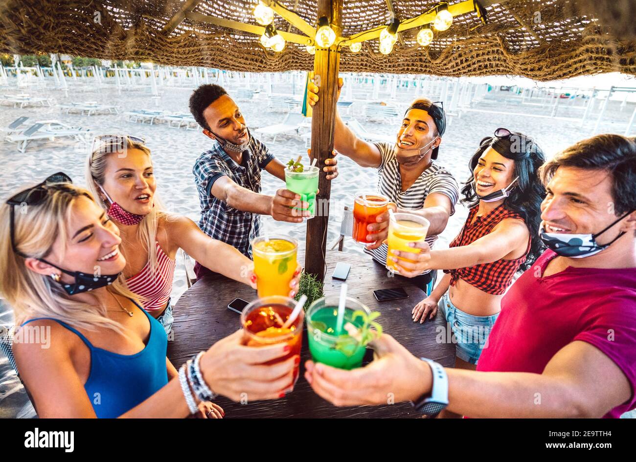 Young trendy friends toasting at beach cocktail bar chiringuito with face mask - New normal summer concept with people having fun together Stock Photo