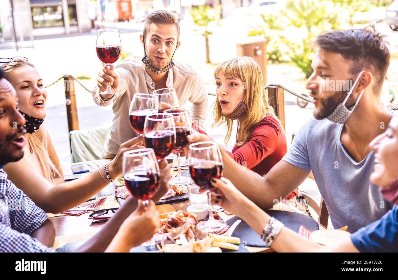 Friends toasting red wine at restaurant bar with face masks - New normal friendship concept with happy people having fun together on sunny day Stock Photo