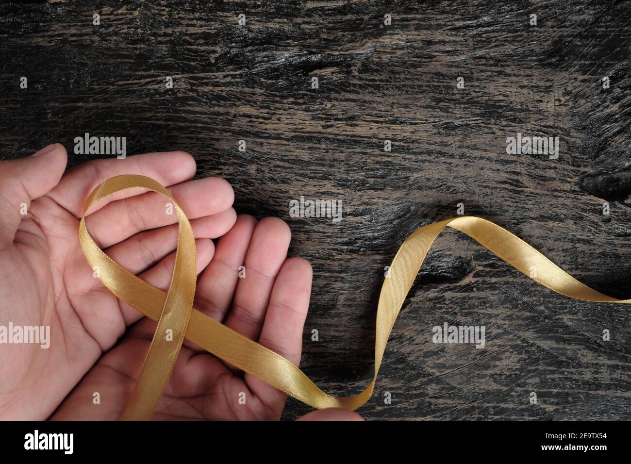Top view of male hand holding gold ribbon on dark wood background. Childhood cancer awareness concept. Stock Photo