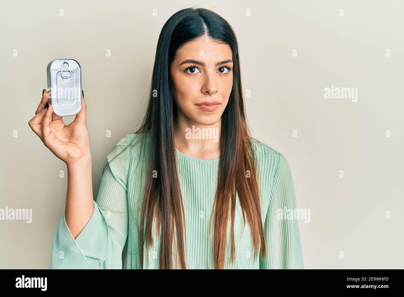 Young hispanic woman holding canned food thinking attitude and sober