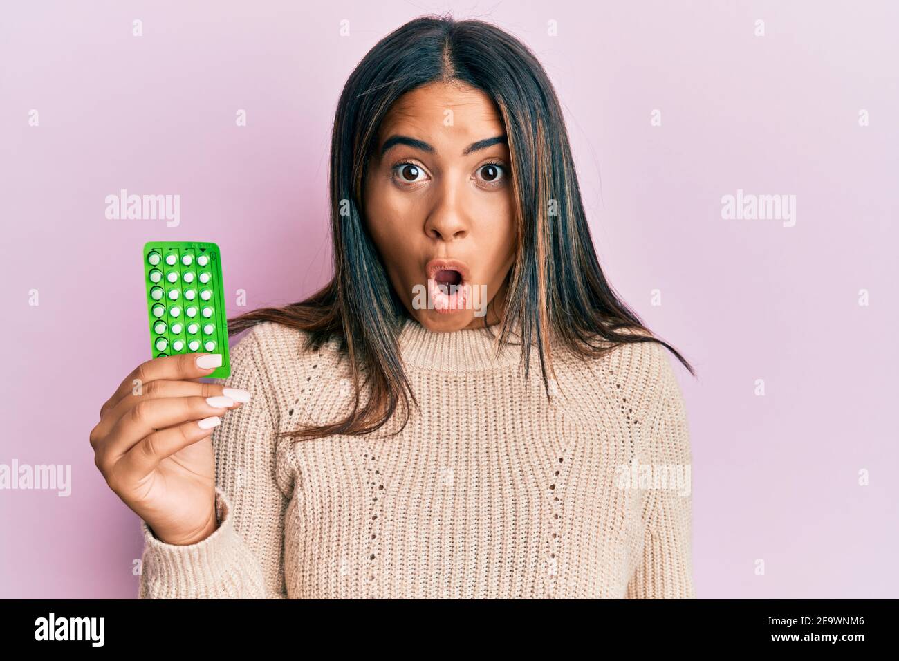 Young latin girl holding birth control pills scared and amazed with