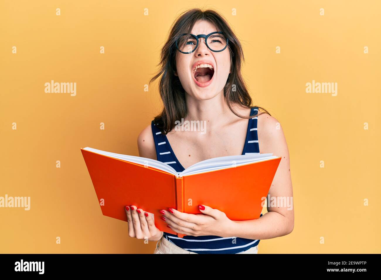 Young beautiful caucasian girl reading a book wearing glasses angry and ...