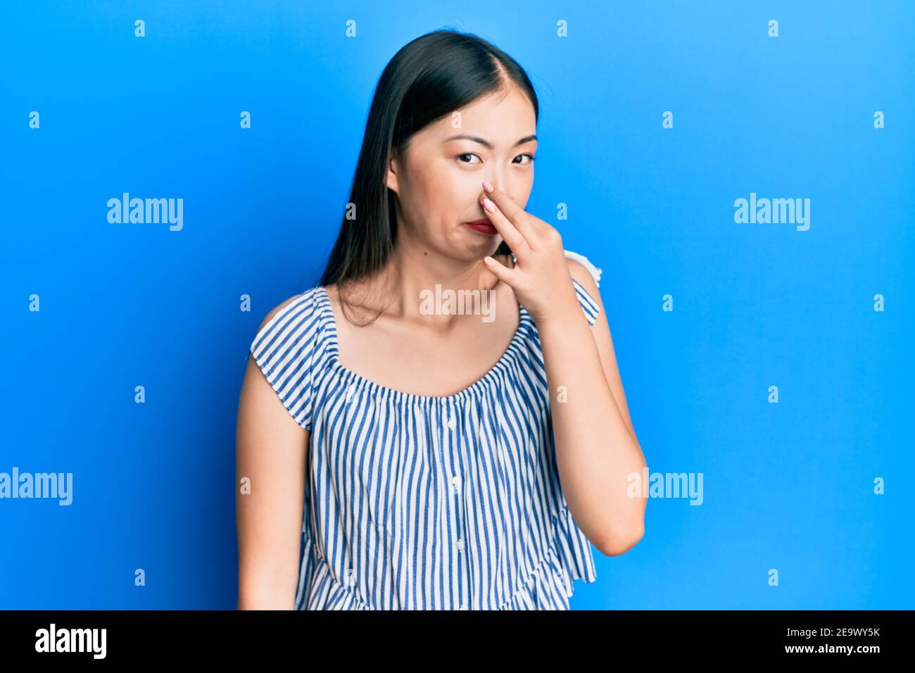 Young chinese woman wearing casual striped t-shirt smelling something stinky and disgusting, intolerable smell, holding breath with fingers on nose. b Stock Photo