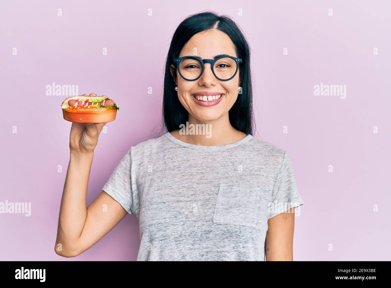 Beautiful young woman eating hotdog looking positive and happy standing and smiling with a confident smile showing teeth Stock Photo