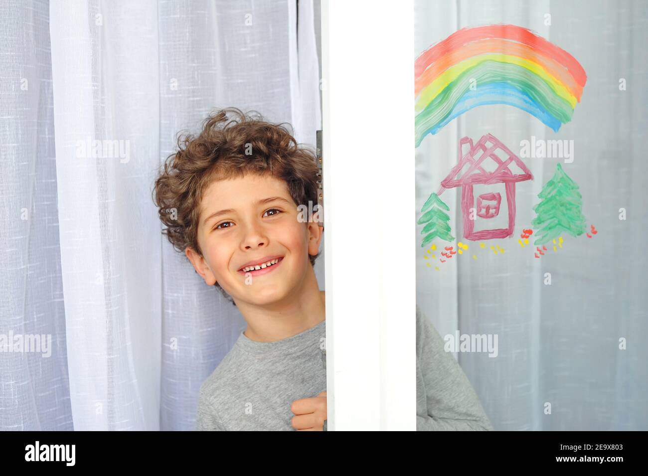 Cute little boy with a rainbow and a house painted on the window during quarantine due to the coronavirus pandemic. Stock Photo