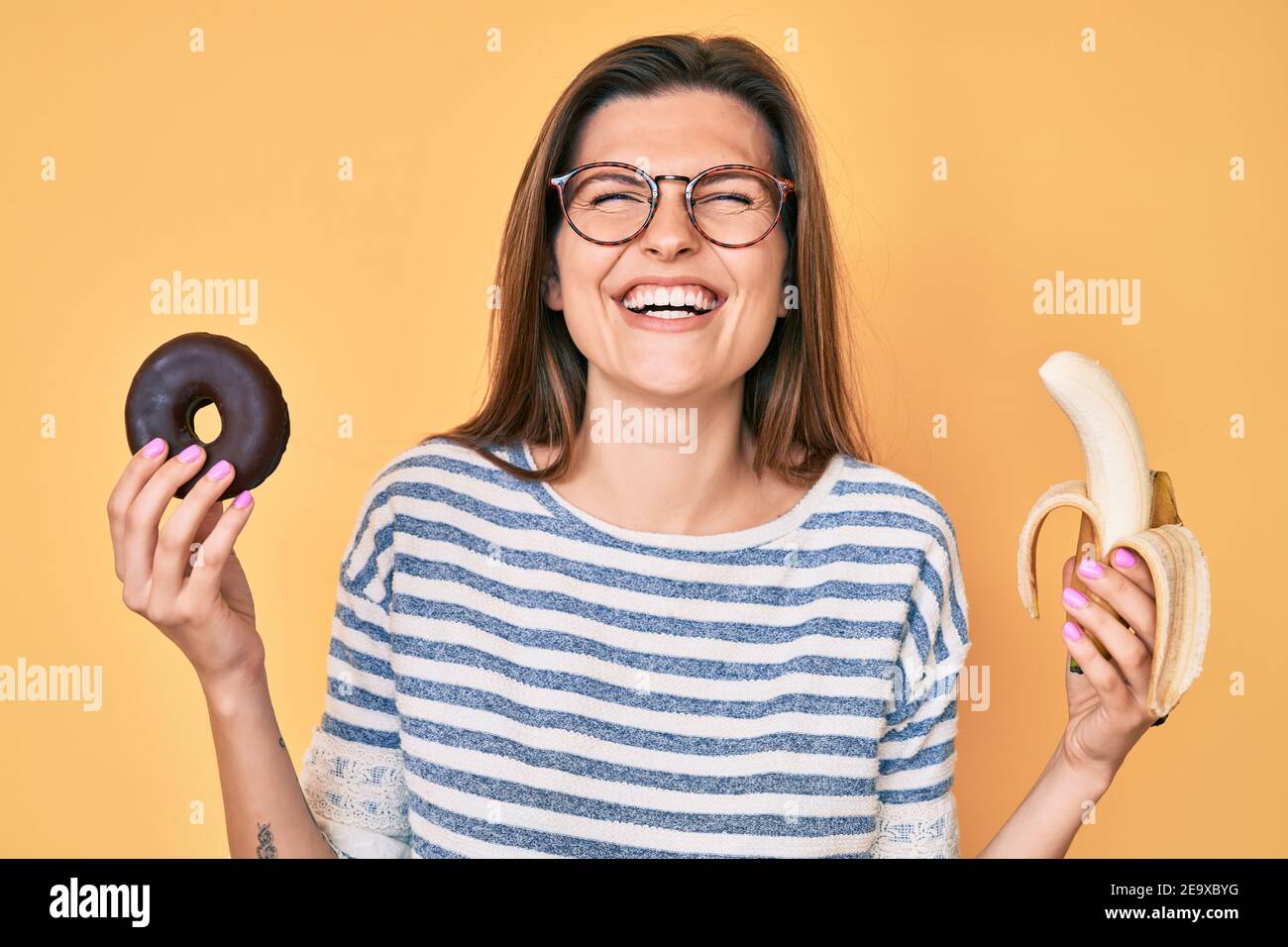 Beautiful caucasian woman holding banana and chocolate doughnut smiling and laughing hard out loud because funny crazy joke. Stock Photo