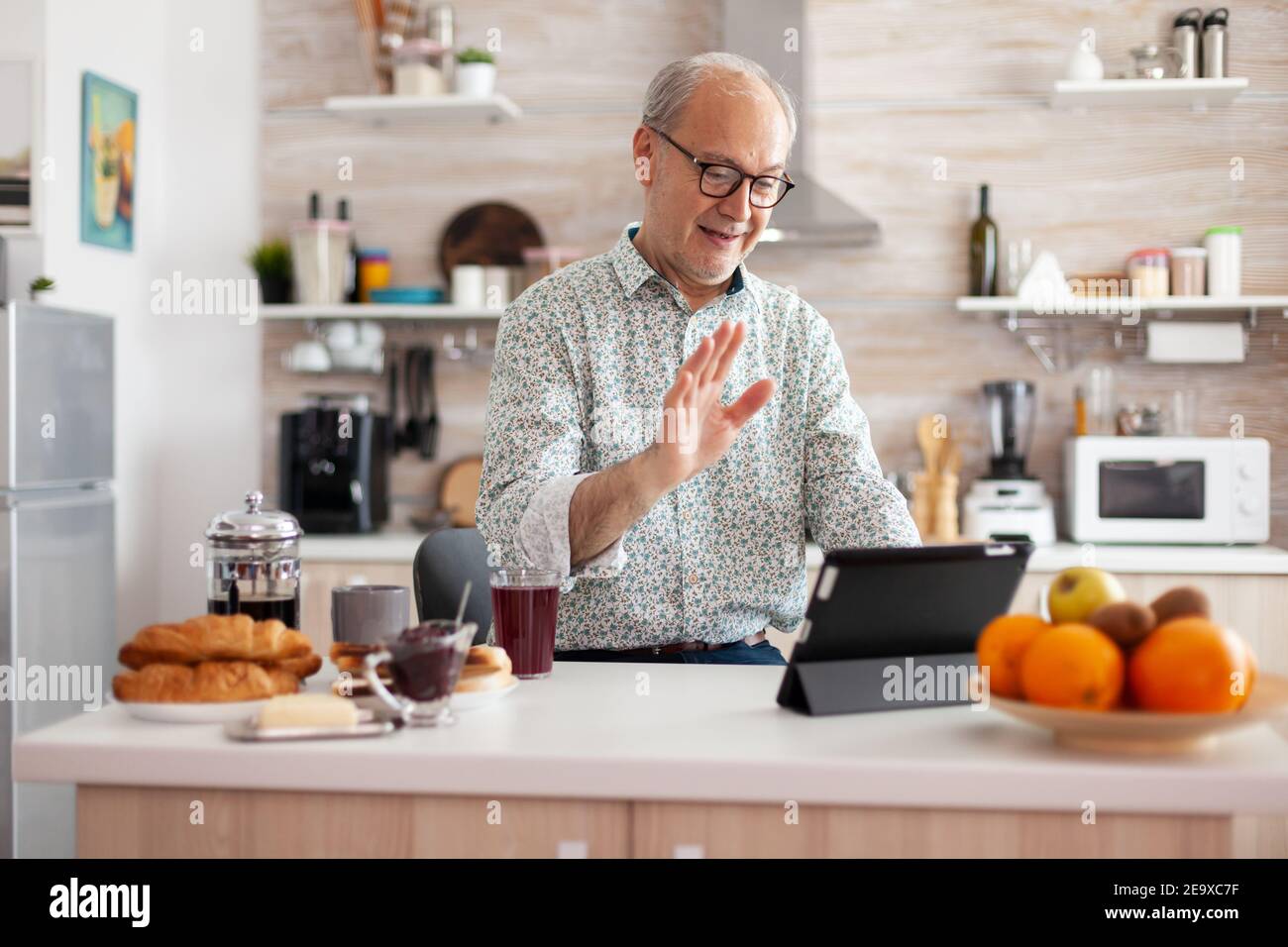 Mature man waving while having a conversation during video call in kitchen enjoying breakfast. Elderly person using internet online chat technology, tablet webcam for virtual conference call Stock Photo