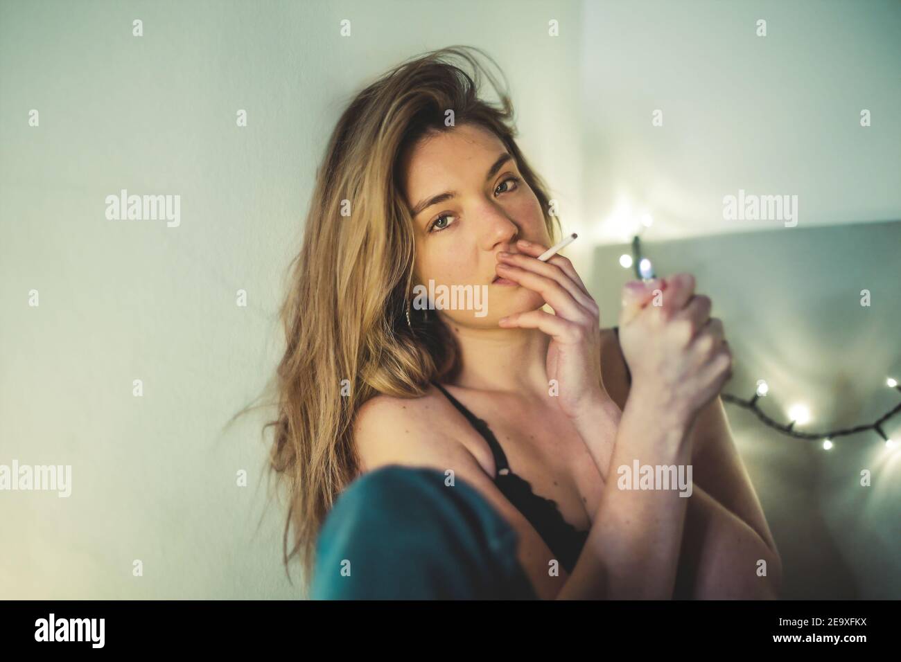 Shallow focus of a woman lighting a cigarette on her bed with light garlands on the background Stock Photo