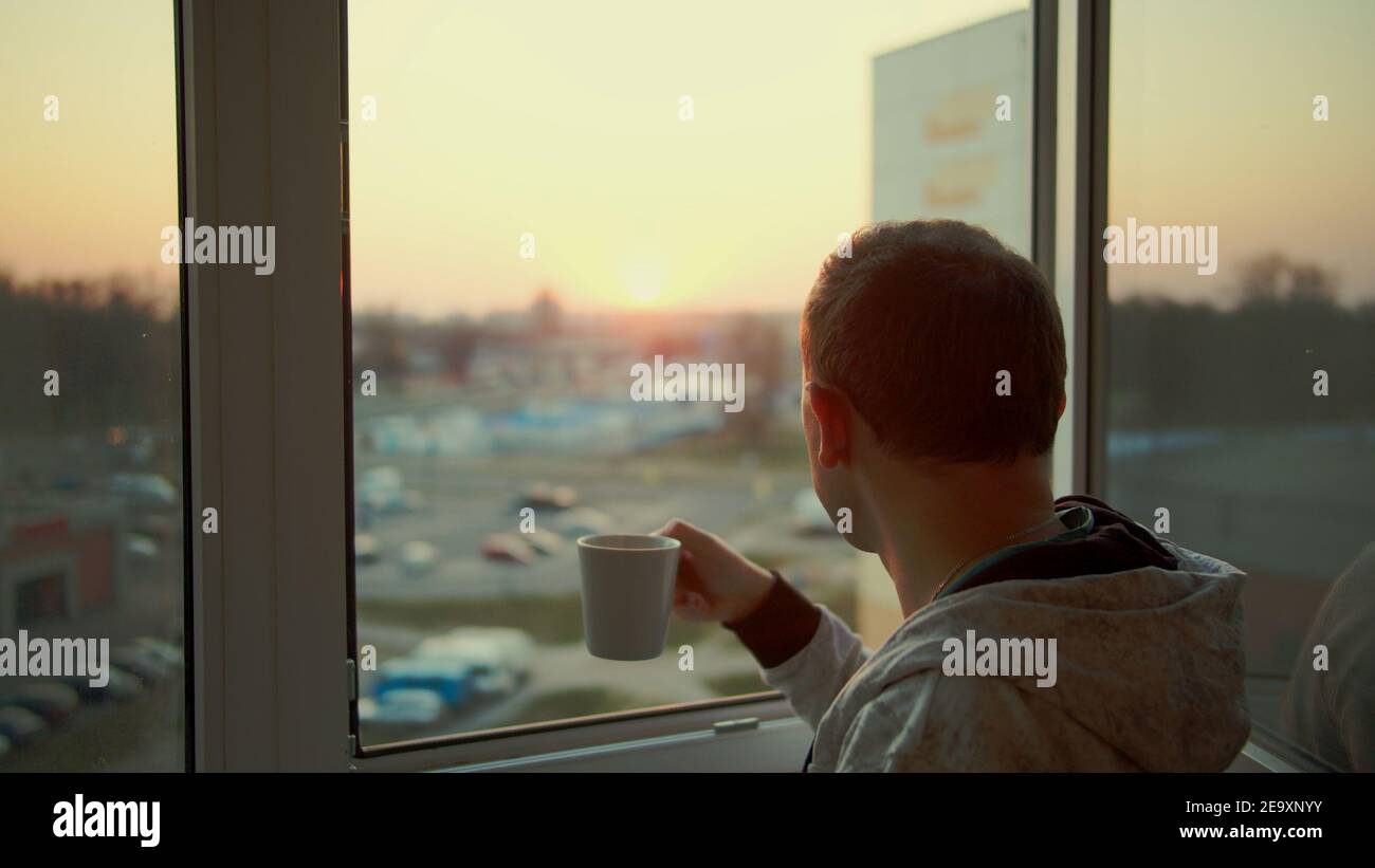 A man stands by the window and watches the sunset, drinks tea from a white mug, rear view Stock Photo
