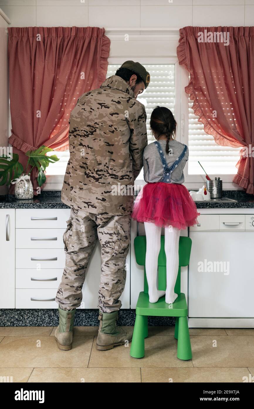 Back view of military father in uniform standing with child in kitchen and washing dishes together Stock Photo