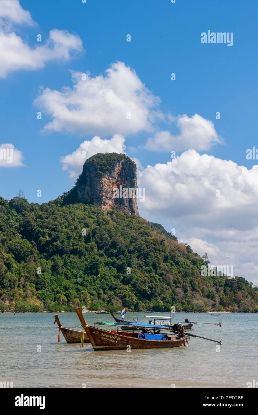 A giant rock formation overshadows the white sand and blue sea and traditional Long Tail boats at Railay Beach, in Krabi, Thailand Stock Photo