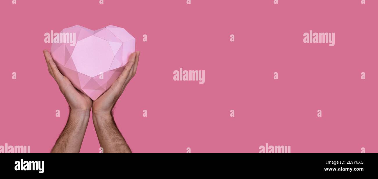 Polygonal three-dimensional pink heart made of paper in the hands of a man. On a pink background. Banner.  Stock Photo