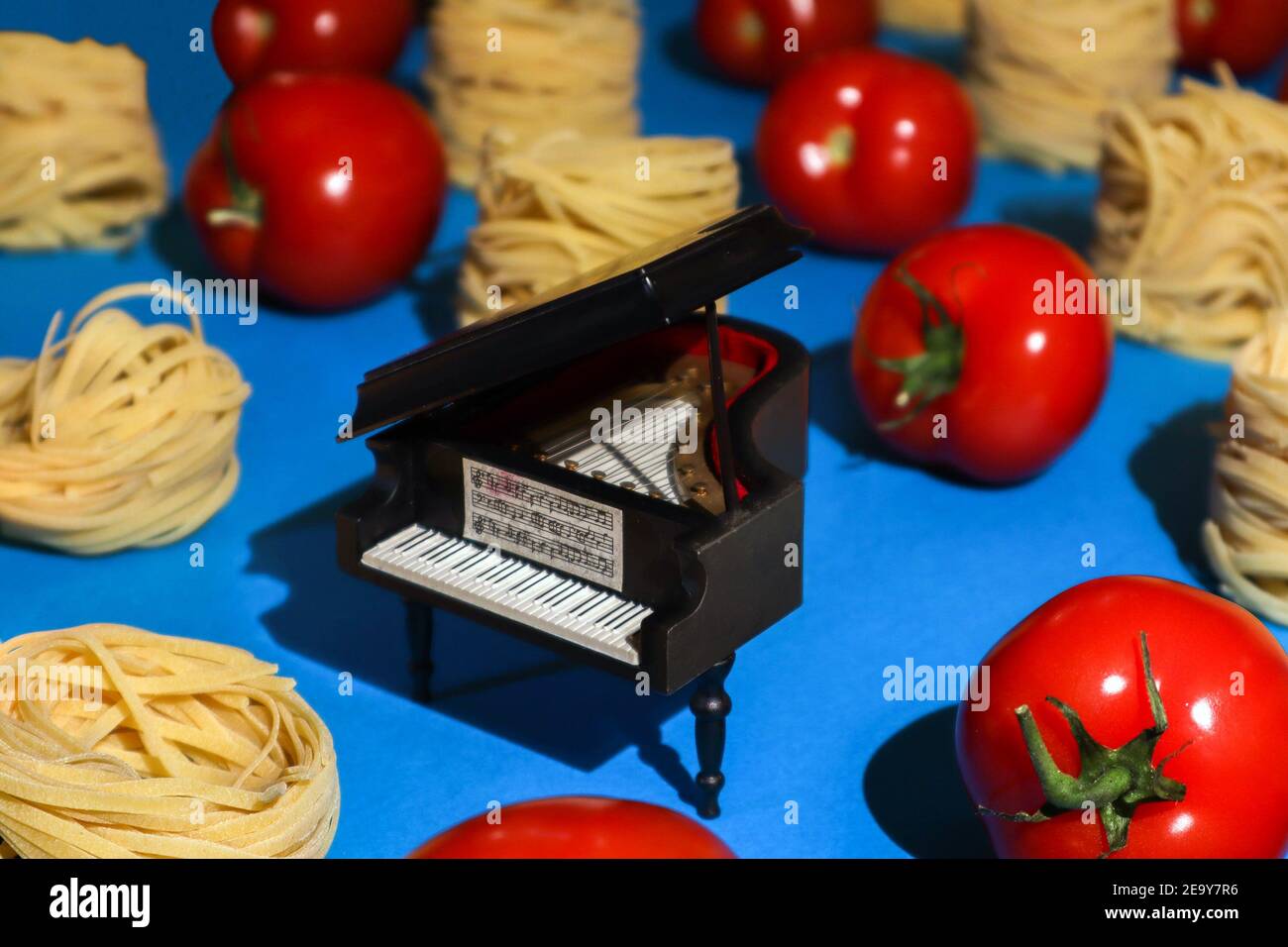 Toy grand piano staying between pasta nests and ripe tomatoes stacked in rows against bright azure background, absurd food photo Stock Photo