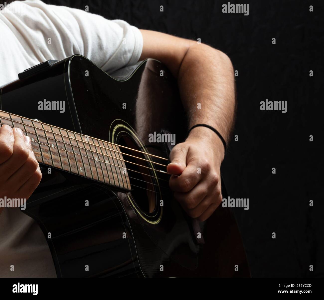 Entertainment concept. Young man playing acoustic guitar, dark background. Guitarist in casual white shirt holds electric black instrument at studio. Stock Photo