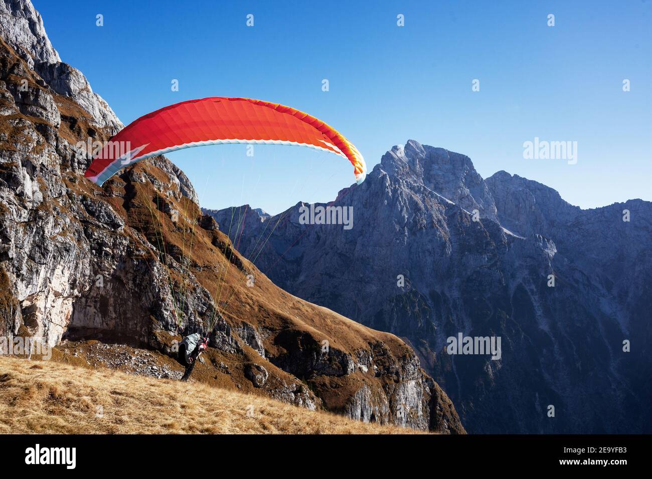 Paraglider with a red parachute taking off from a mountain meadow among the surrounding mountains with Jalovec in the foreground. Stock Photo