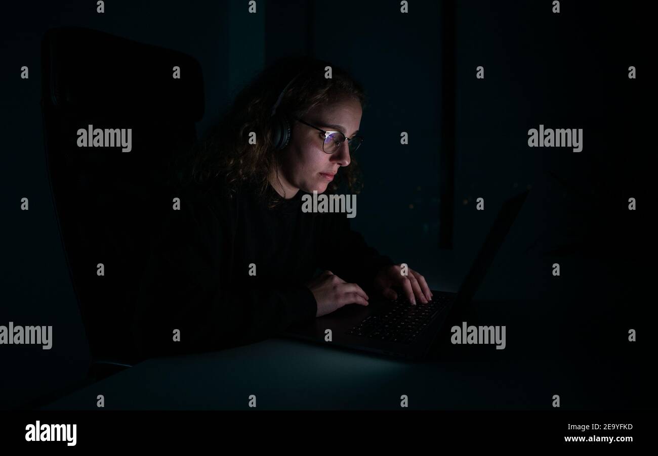 Woman with headphones typing on her laptop in the dark of the night ...