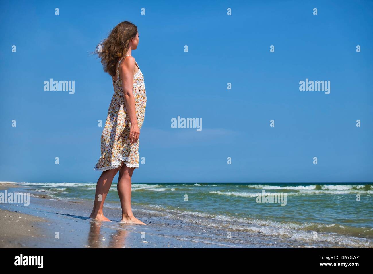 Young girl in a flower sundress stands on the seashore and looks into the distance Stock Photo
