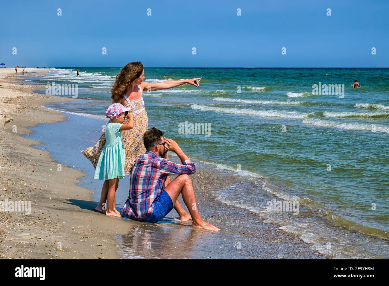 Girl shows something far into the sea to her husband and daughter. Young happy family walking along the seashore Stock Photo