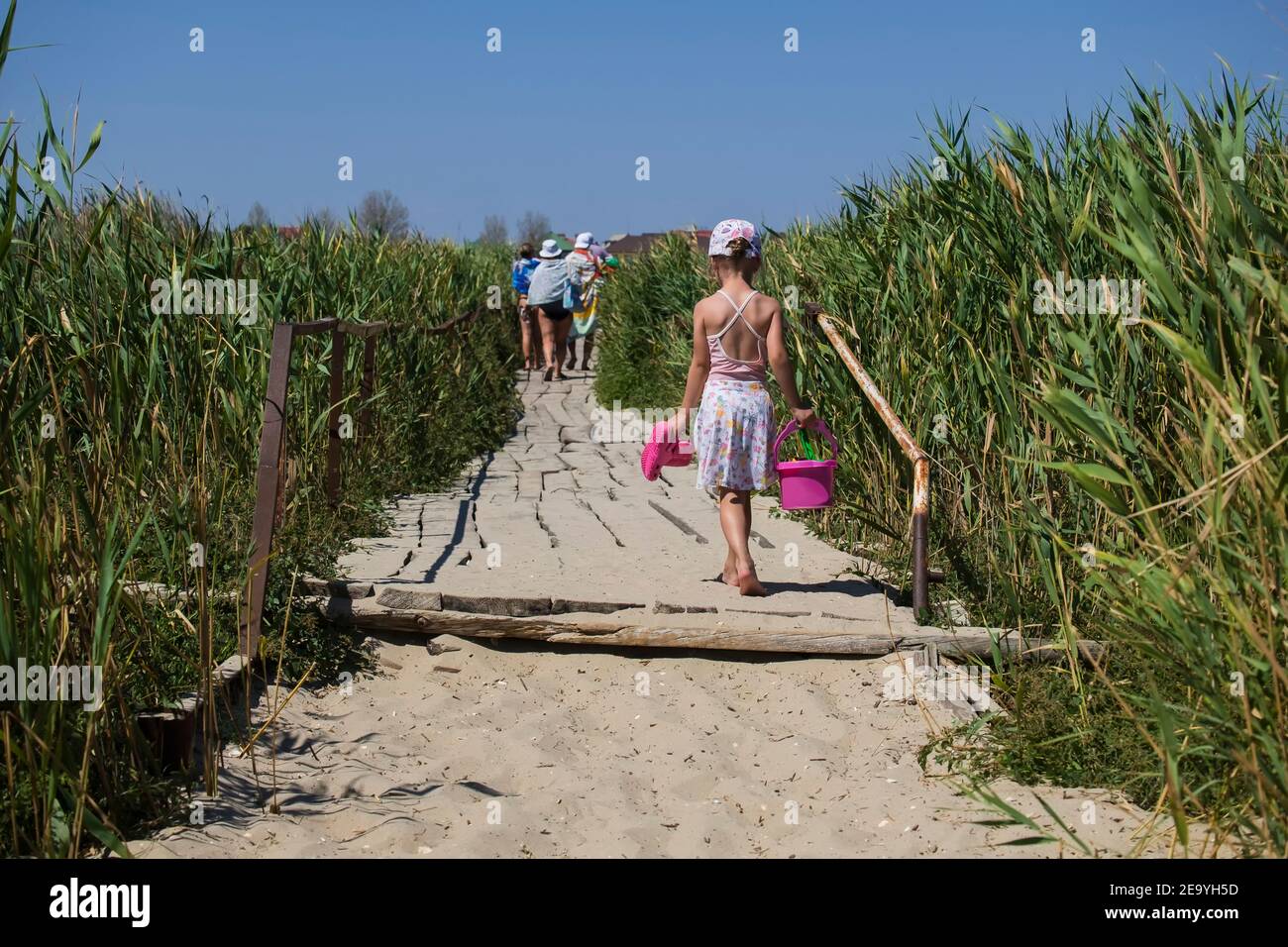 Small beach girl walks over a wooden bridge with a bucket and slippers in her hands Stock Photo