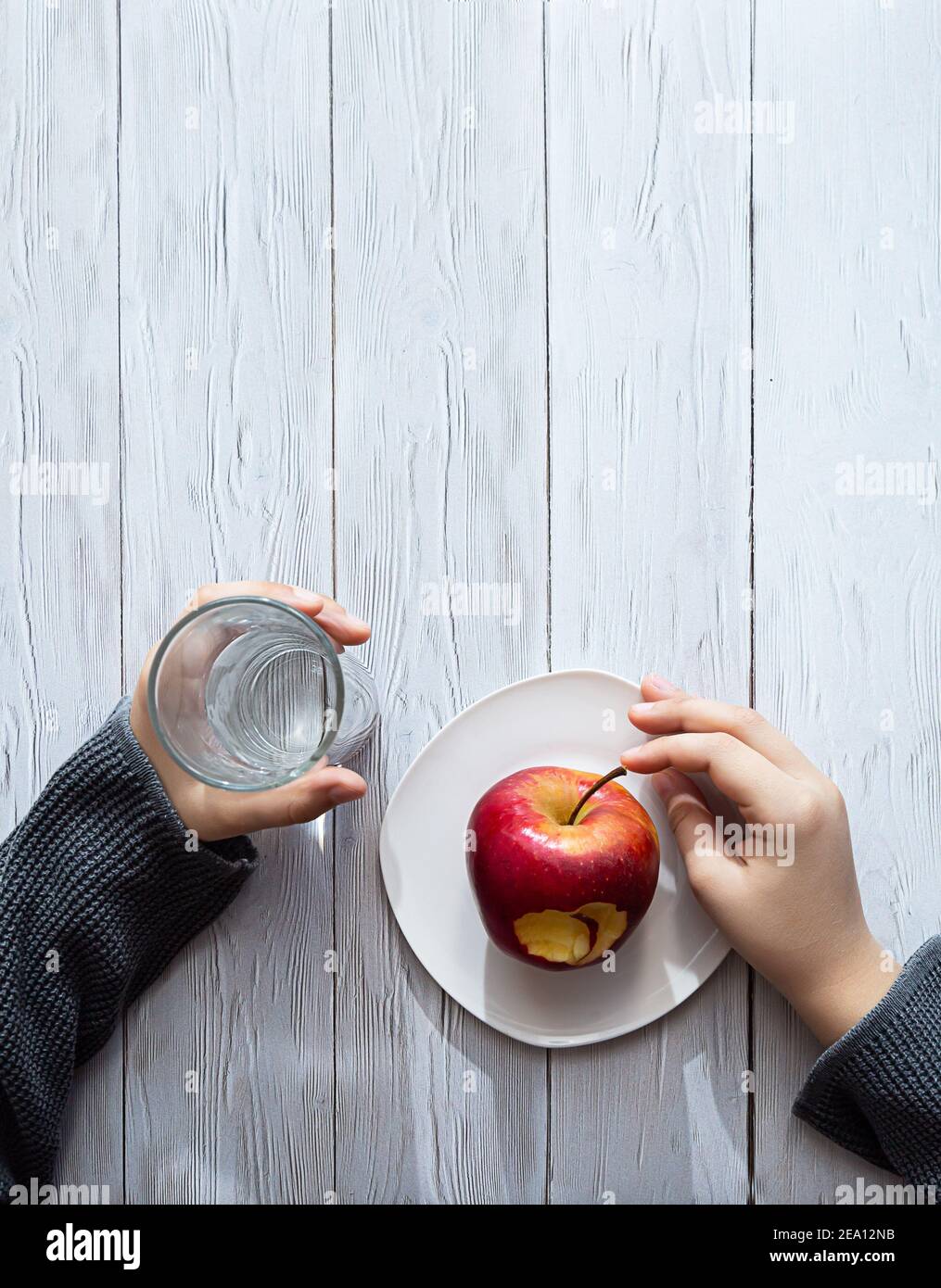 Minimal healthy snack concept. The child's hands are holding an apple and a glass of water. Still life on a light wooden table with shadows and highli Stock Photo