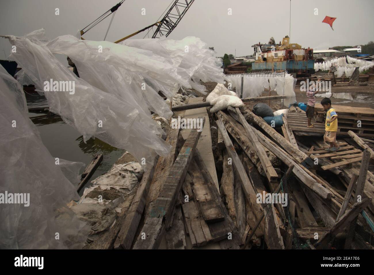 Children playing kite close to wooden materials, coastal protection structure, plastic covers being dried in the sun and heavy machinery in the coastline of Jakarta, Indonesia. Stock Photo