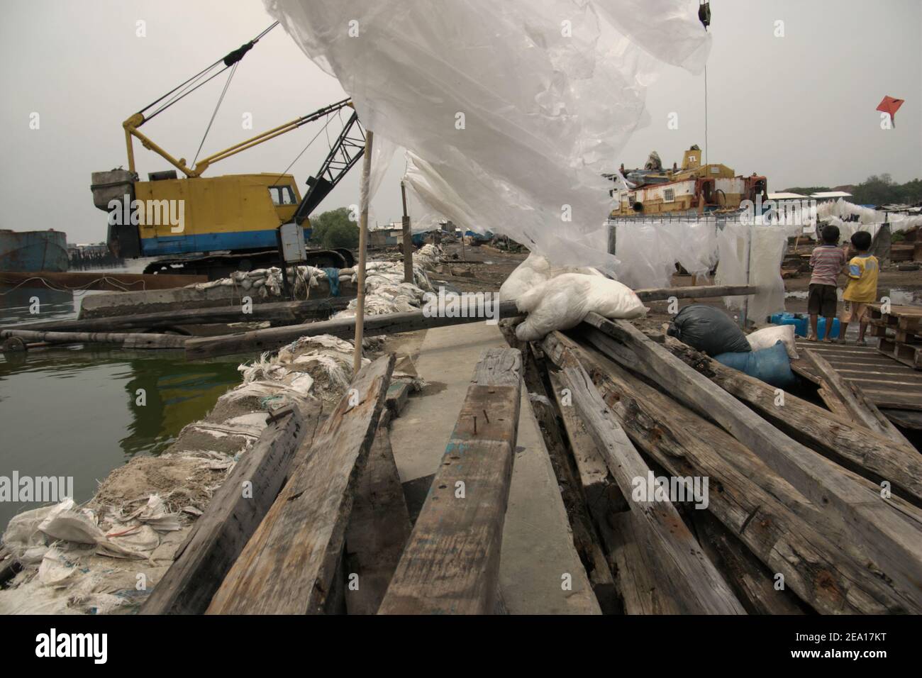 Children playing kite close to wooden materials, coastal protection structure, plastic covers being dried in the sun and heavy machinery in the coastline of Jakarta, Indonesia. Stock Photo