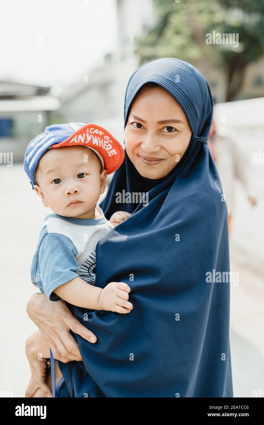 Thai Muslim woman wearing a blue hijab smiling at the camera, holding a baby with a cap, looking at the camera, with sunny bright background. Stock Photo