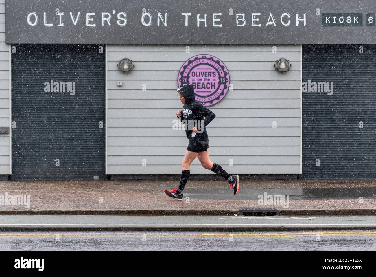 Southend on Sea, Essex, UK. 7th Feb, 2021. Storm Darcy reached Southend on Sea later than initially forecast, with the area remaining wet overnight. Snow and sleety flurries arrived from around 7am, which didn’t deter joggers. A female runner is passing Oliver's on the Beach restaurant kiosk. Owned by members of Jamie Oliver's family. COVID 19 lockdown warnings remain in place Stock Photo