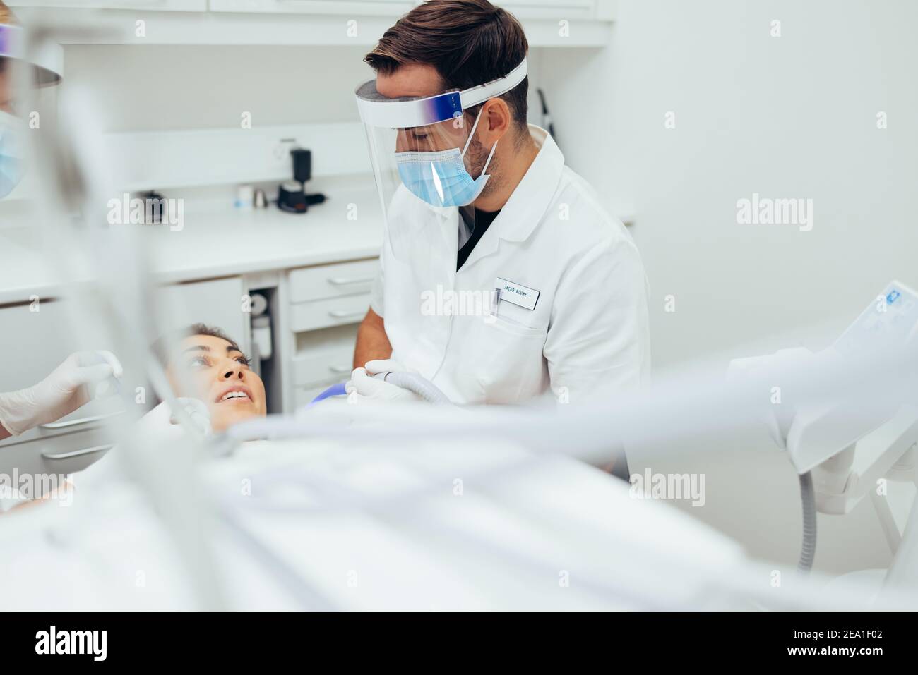 Dentist treating a female patient at dental clinic. Male doctor wearing  mask and face shield discussing treatment with patient at dental clinic. Stock Photo