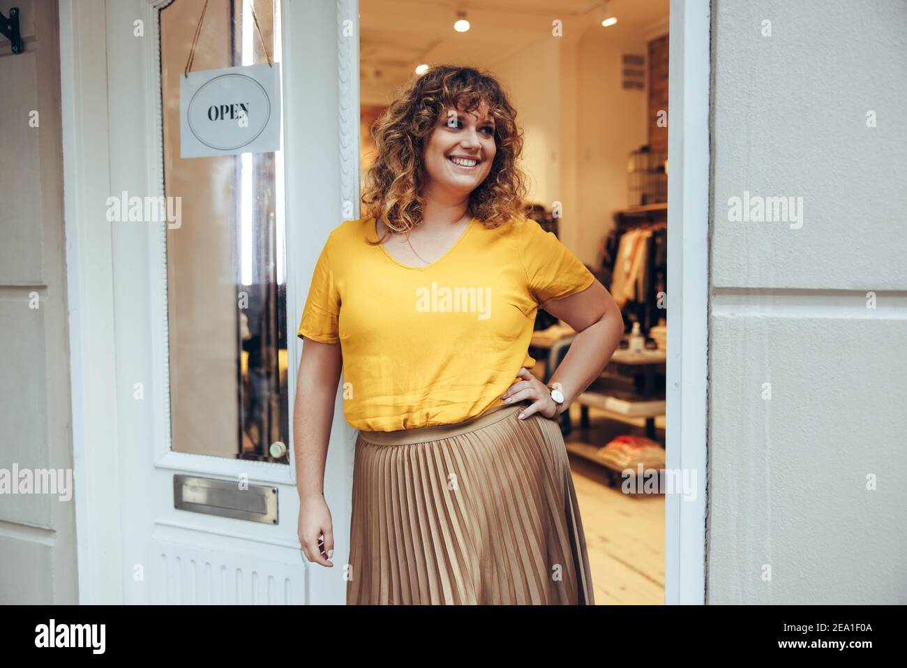 Portrait of a happy mid adult woman standing with hand on hips at fashion clothing store doorway. Small business owner standing at store entrance with Stock Photo