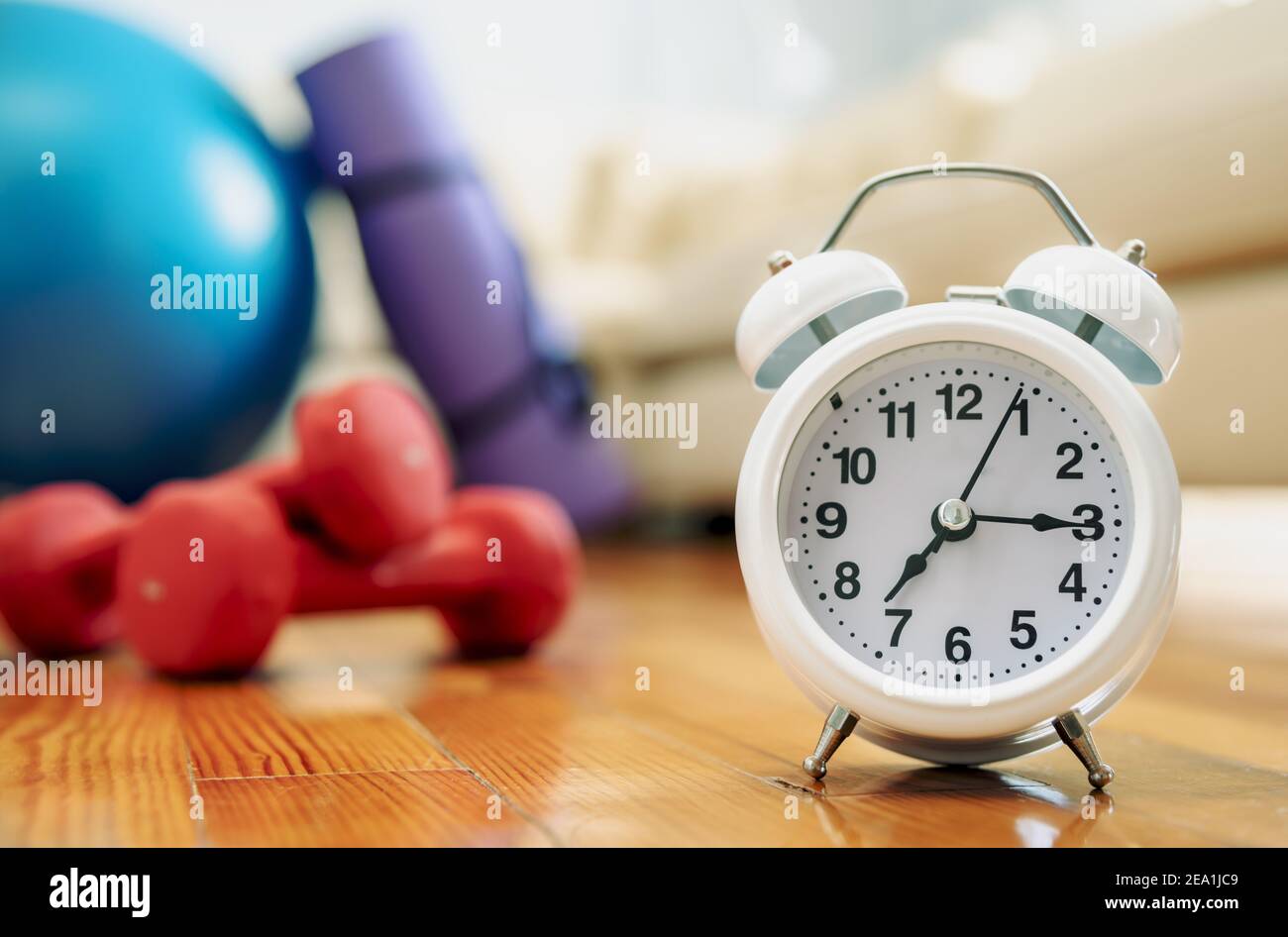 Alarm clock on the wooden floor with unfocus sports equipment and couch on background. Time for exercising concept Stock Photo