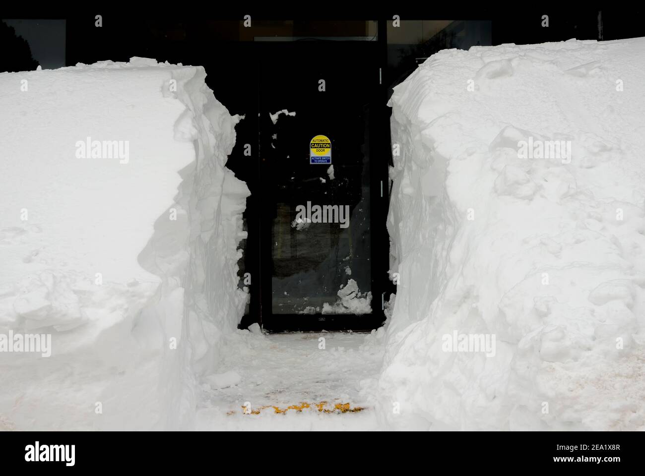 A path cut through a snowbank leading to a door. The sign on the door says CAUTION AUTOMATIC DOOR ACTIVATE SWITCH TO OPERATE. Stock Photo