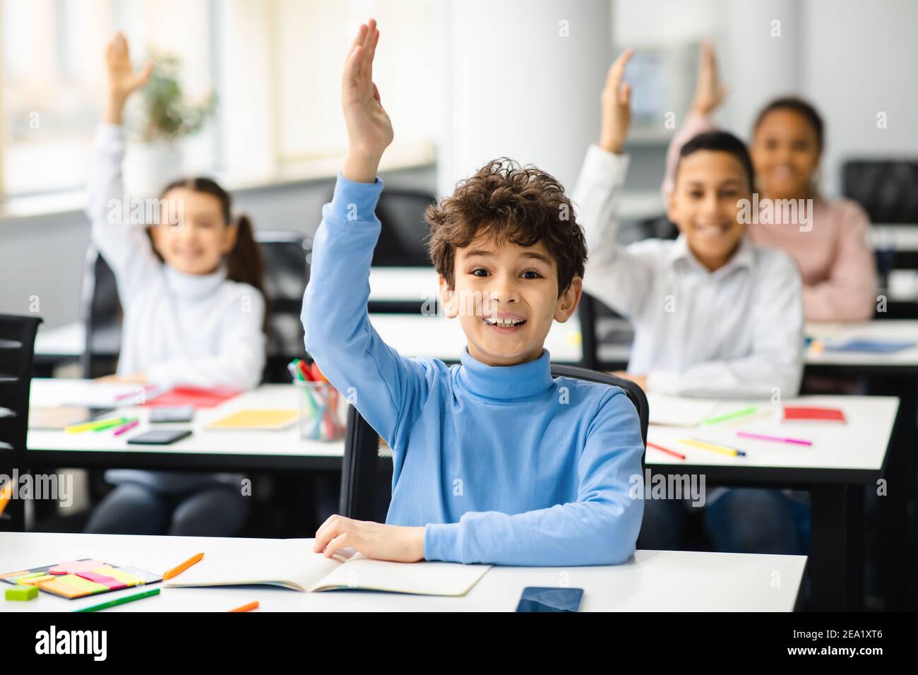 Diverse small schoolkids raising hands at classroom Stock Photo