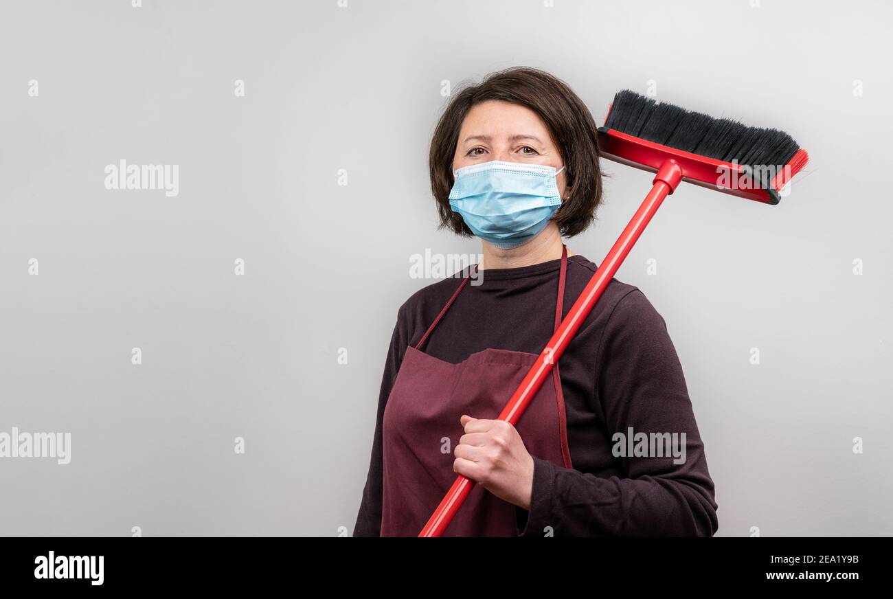 Woman holding a broomstick over her shoulder wearing an apron and a mask. Isolation during the Covid pandemic. Stock Photo