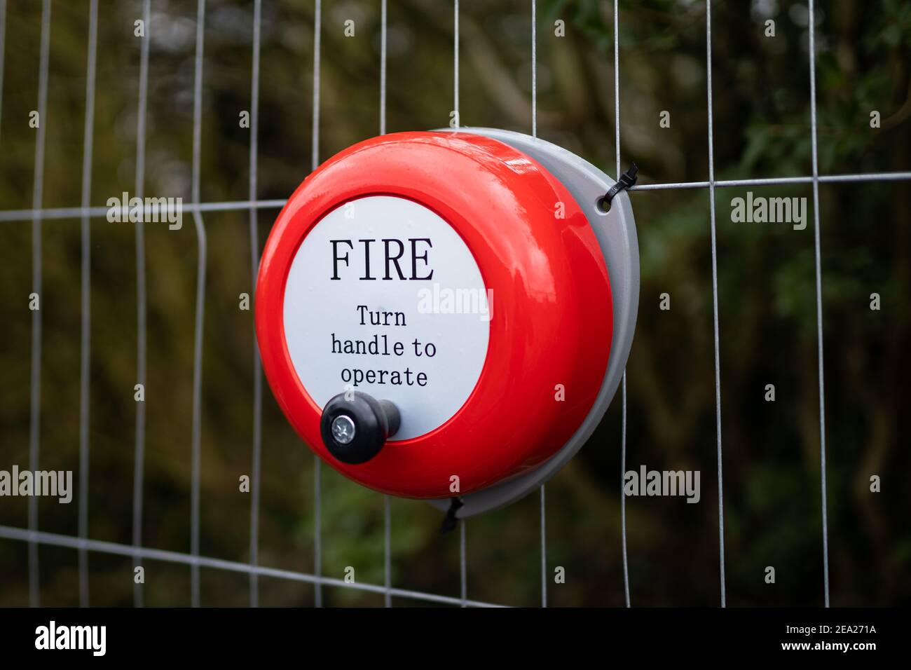 Bright red metal fire alarm bell turn handle to operate alert manual steel ringing on construction building site attached to metal fence Stock Photo