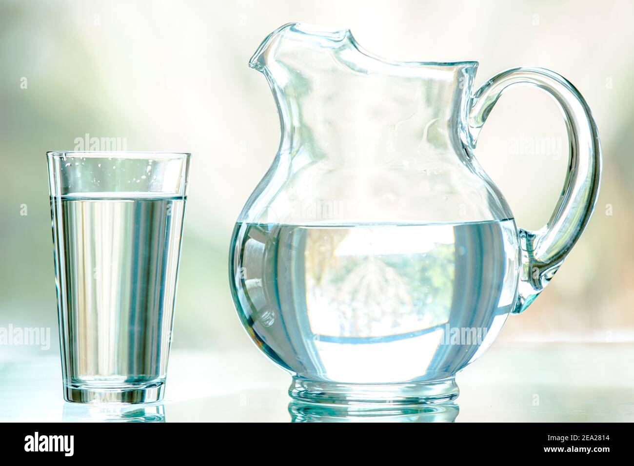 A clear pitcher and glass filled with water. It is shot straight on on a glass table, with a nature background seen through a window .Shallow DOF. Stock Photo