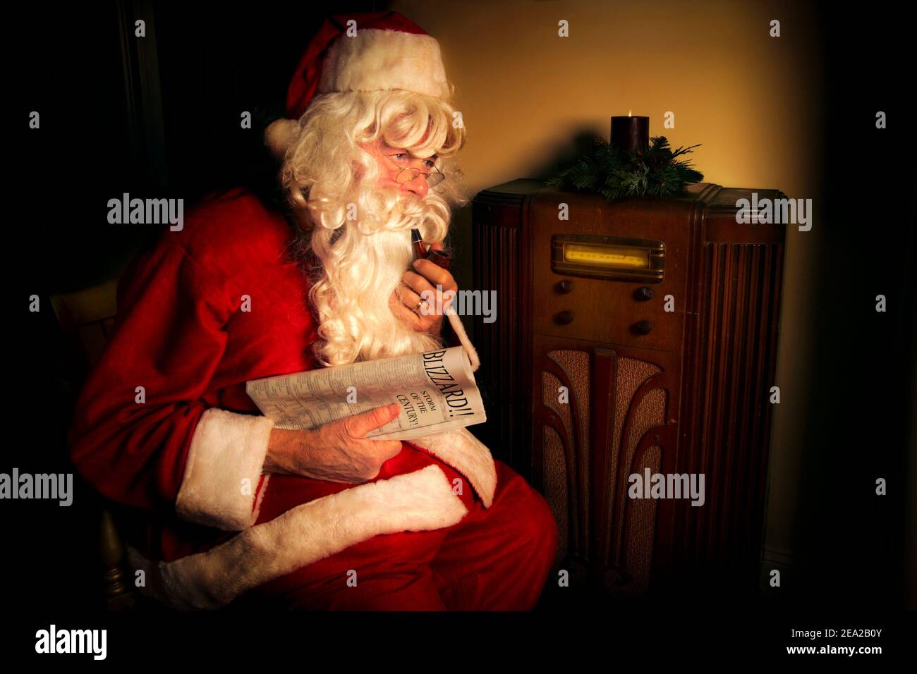 Santa Claus listens to the weather report an old fashioned radio. He’s holding a newspaper that says “Blizzard!” on it. Stock Photo