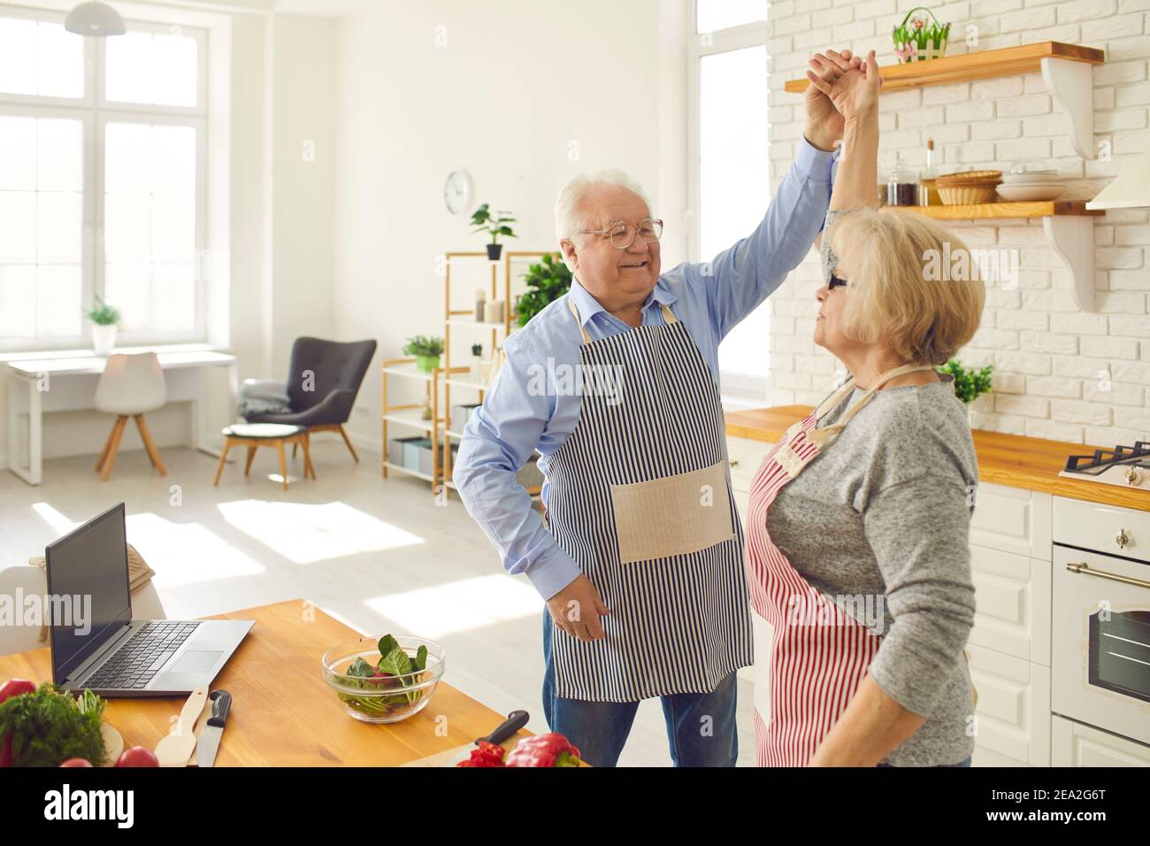 Happy senior woman dancing with mature loving husband in the kitchen while cooking dinner. Stock Photo