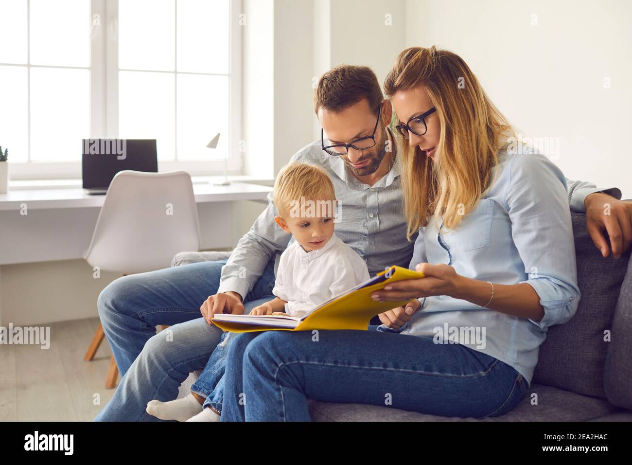 Mom, dad and son read a book of stories together sitting on the couch in a bright room at home. Stock Photo