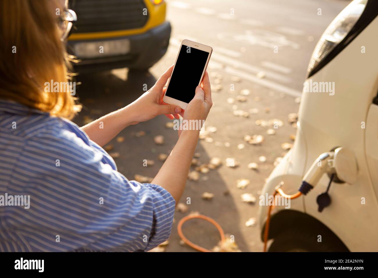Hands with smartphone on a background of rental car at the charging station for electric vehicles. Car sharing.  Stock Photo