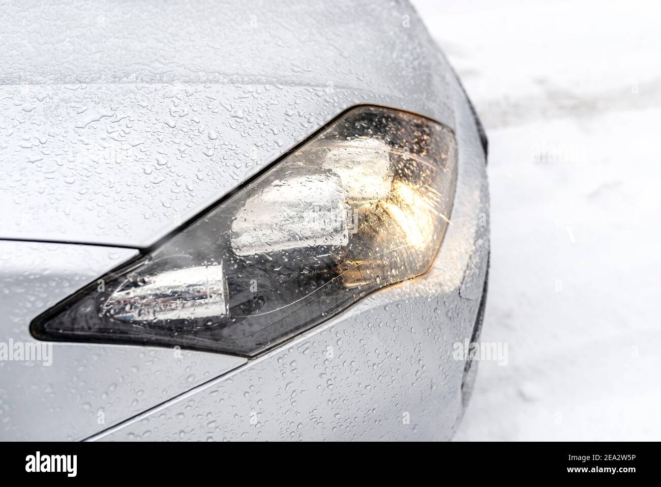 The dipped headlights on a silver car standing on a snowy road in the forest, visible snow falling. Stock Photo