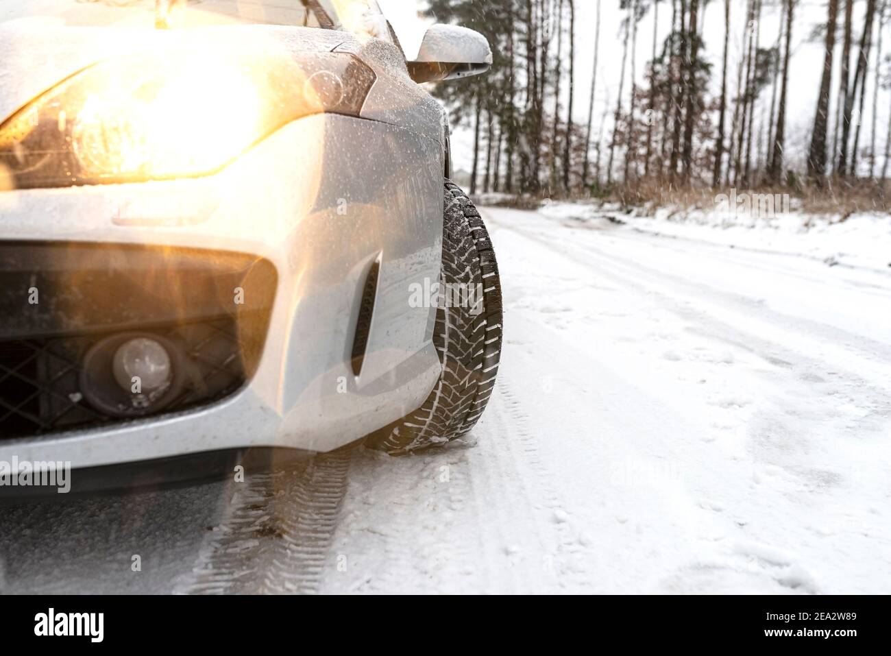 The dipped headlights on a silver car standing on a snowy road in the forest, visible new winter tire and falling snow. Stock Photo
