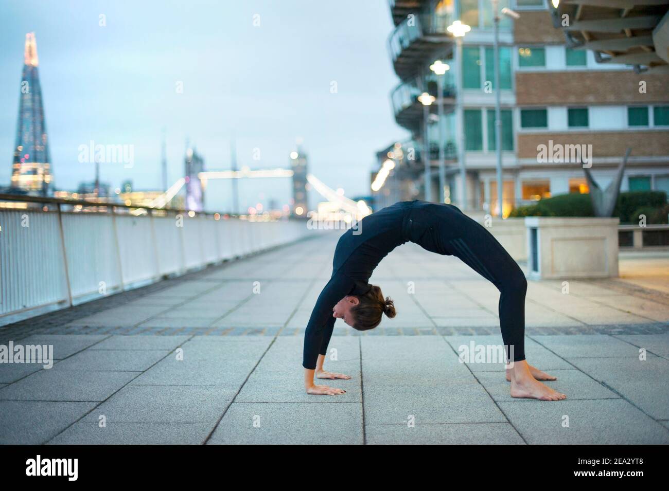 Flexible young adult female doing bridge pose on street promenade, along the Thames with Tower Bridge and the Shard in the background. London, UK Stock Photo